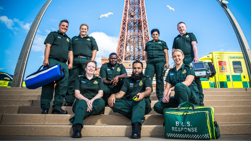 Ambulance crew mates in uniform sitting on steps on Blackpool prom with Blackpool Tower in the background and a response kit bag to the right.