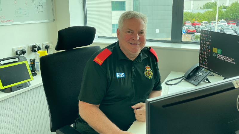 Chief Consultant Paramedic Mike Jackson in green uniform sat at desk with computer with window and car park in background.