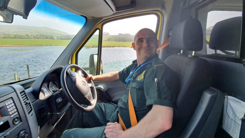 Paramedic Maciej in the cab of a stationary ambulance pulled over at the side of a lake.