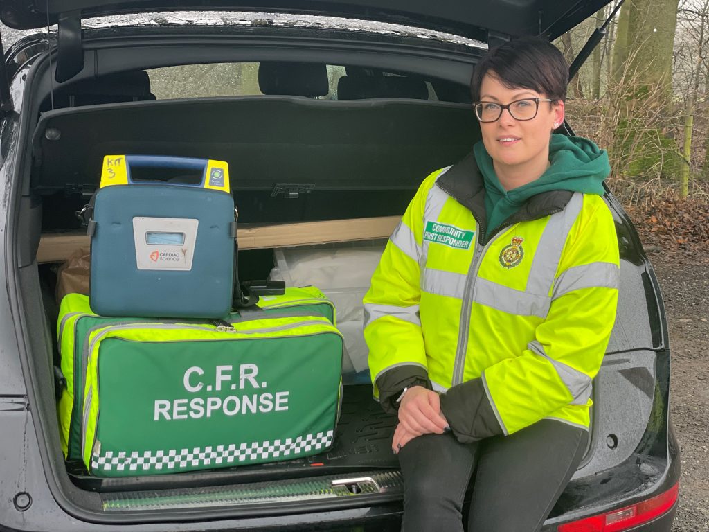 Female Community First Responder smiling from back of their car with defibrillator and medical response bag