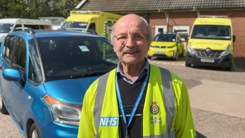 Volunteer car Driver Colin standing in front of his car in his uniform smiling.