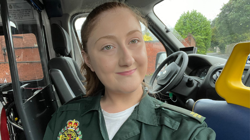 Ambulance care assistant Kaitlyn in uniform smiling to camera in the front of a patient transport service ambulance.