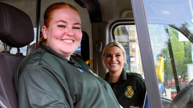 Two women smiling sat a cab of an ambulance thw woman on the left has red hair the has blonde hair.