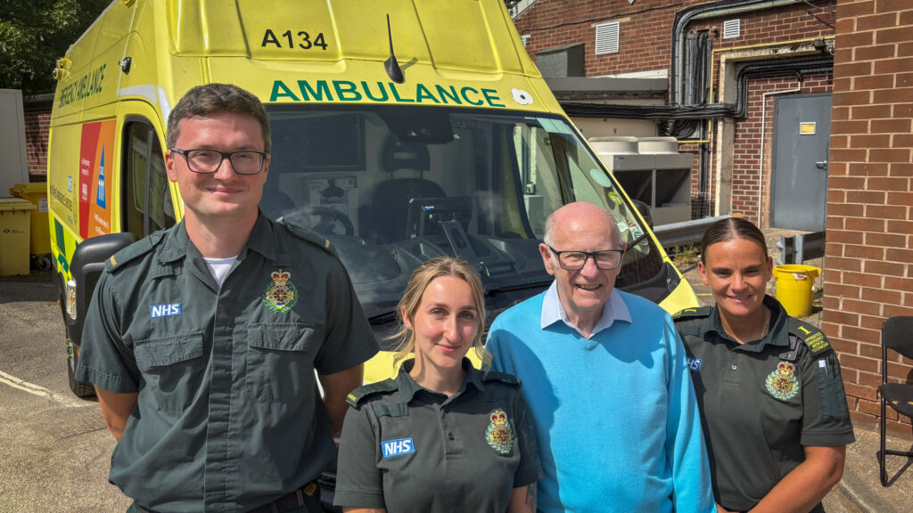 A elderly man in blue jumper (Alan) stood in between three paramedics: two woman one man wearing green uniform. They're all stood in front of an ambulance on a sunny day.
