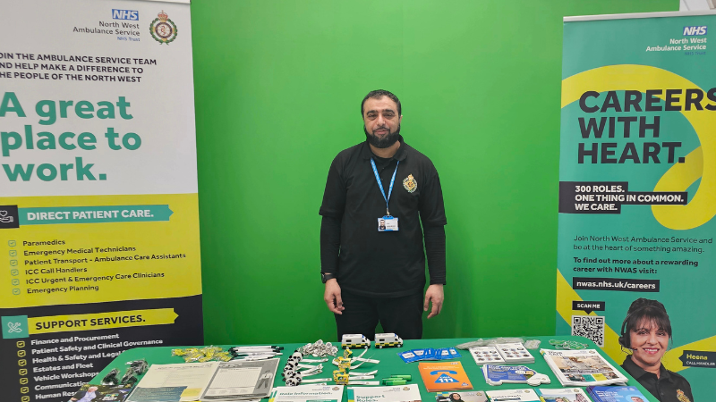 Jaffa in NWAS uniform standing at a NWAS careers fair stand with various banners and promotional materials.