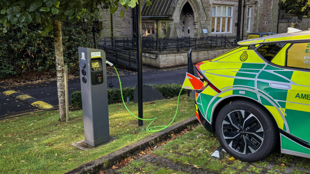 ambulance response vehicle plugged into an electric car charger in a car park in front large old looking house.