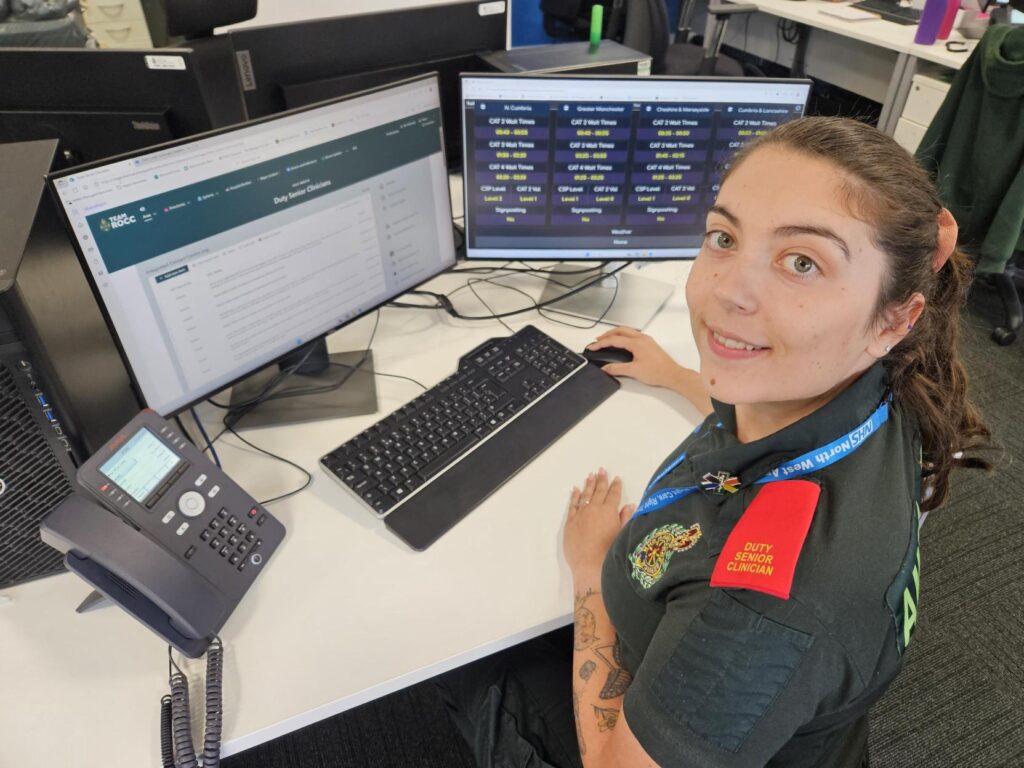 Duty Senior Clinician Asha in green NWAS uniform and red epaulettes, sat smiling to camera in front of control centre monitor screens.