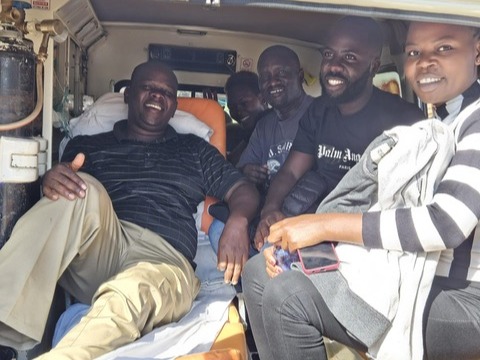 A group of people smiling inside a vehicle, one lying on a stretcher.