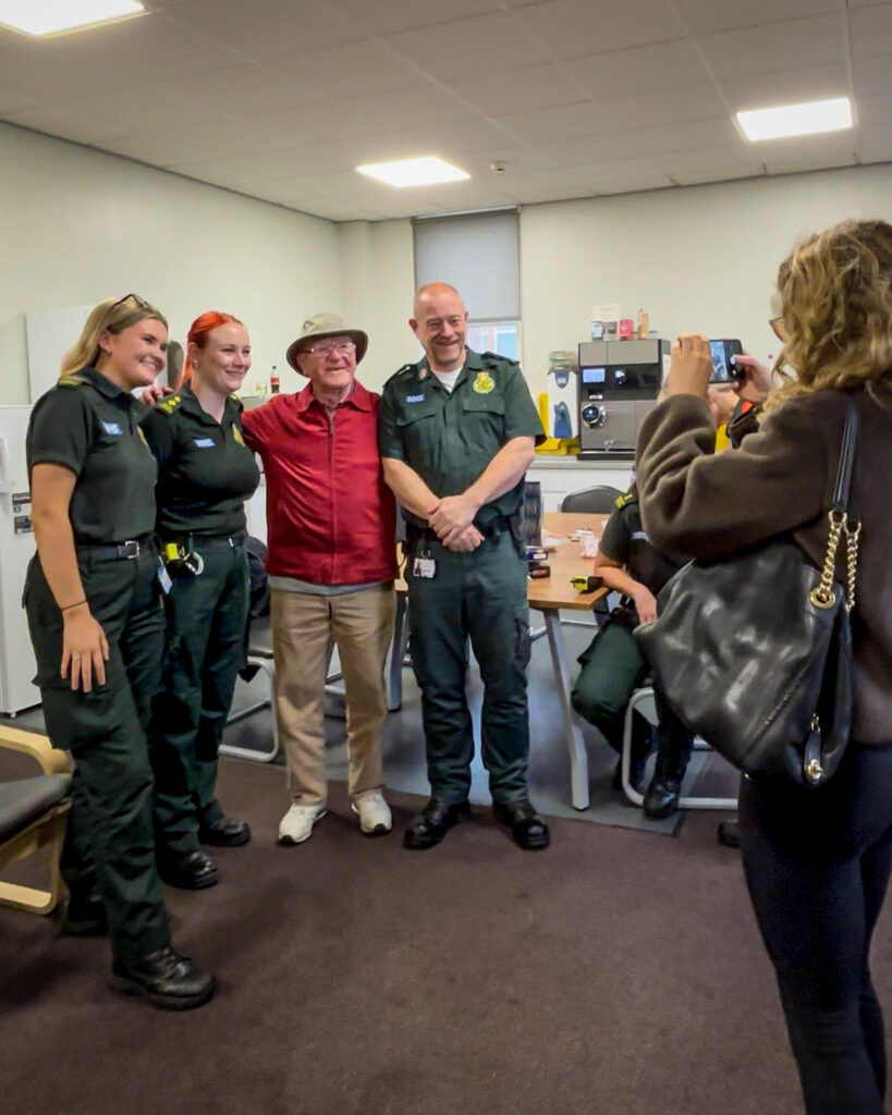 Three paramedics and another individual in a red shirt and hat smiling for a photo taken by a person with a phone in an office setting.