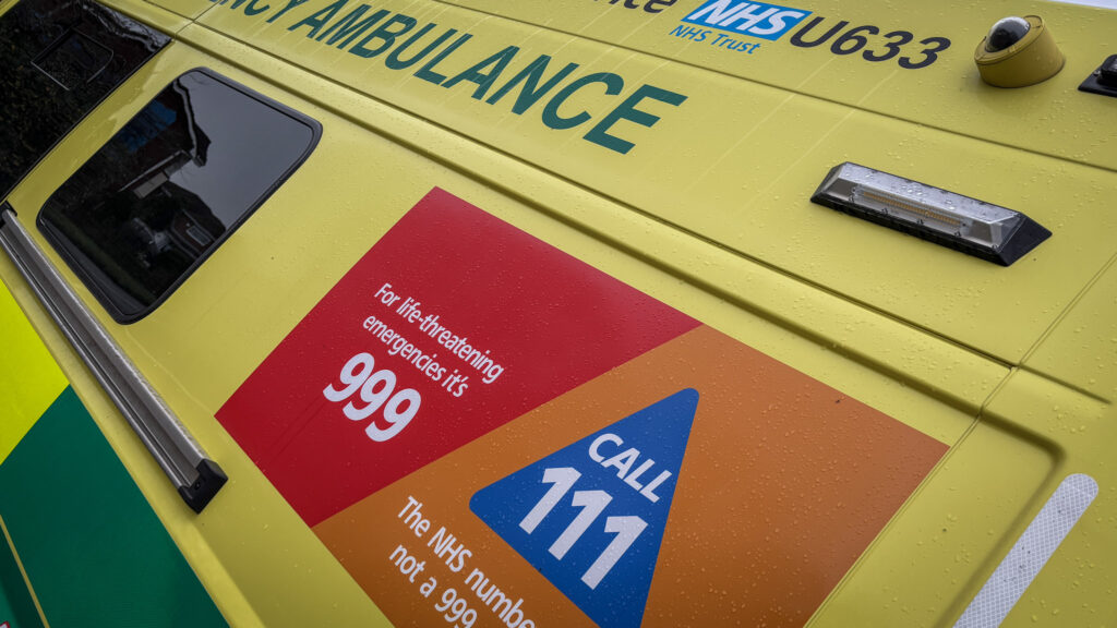 Close-up of an NHS ambulance sign with instructions: "For life-threatening emergencies CALL 999" and "The NHS number when it's not a 999 emergency CALL 111."
