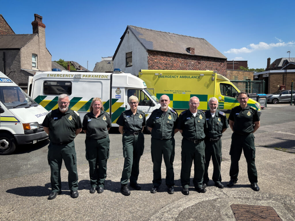 Seven people stood in front of a row of a mixture of modern and vintage ambulances