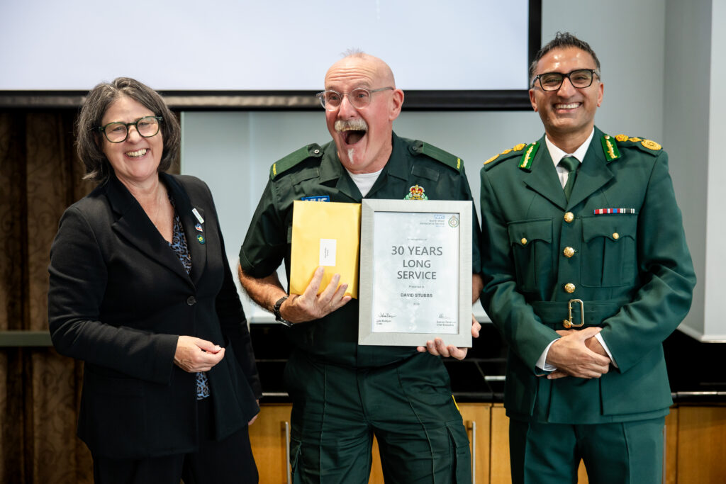 three people on a stage, the middle person is excitedly smiling holding a 30 years service certificate