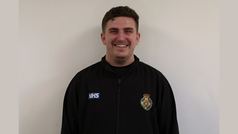 Rhys wearing a black top with the NHS crest on and North West Ambulance Service Crown Badge.