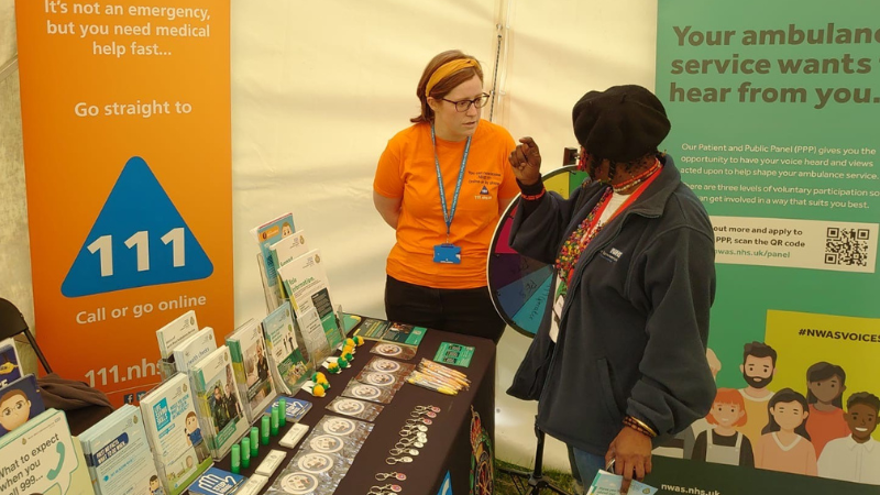 Two individuals engaging in a conversation at a medical service awareness booth. A prominent triangular blue sign reads "111" on one banner. Additionally, a poster highlights voluntary participation in NHS with a QR code and website link for more information.