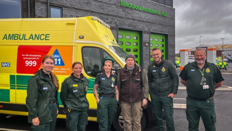 Five paramedics in uniform standing in front of an NHS ambulance at the Blackpool Ambulance Hub. Standing in the middle is the patient, Chris Jameson.