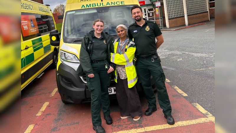 Three NWAS staff stood at the front of an ambulance, two are wearing green uniforms and the middle person is in a high vis jacket over plain clothes.