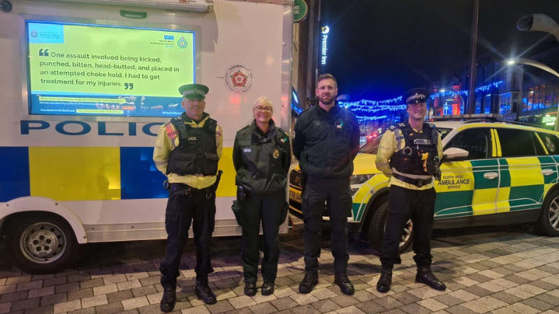 Four emergency service workers, including police officers and ambulance service staff, standing in front of their vehicles – a police car and an ambulance – at night