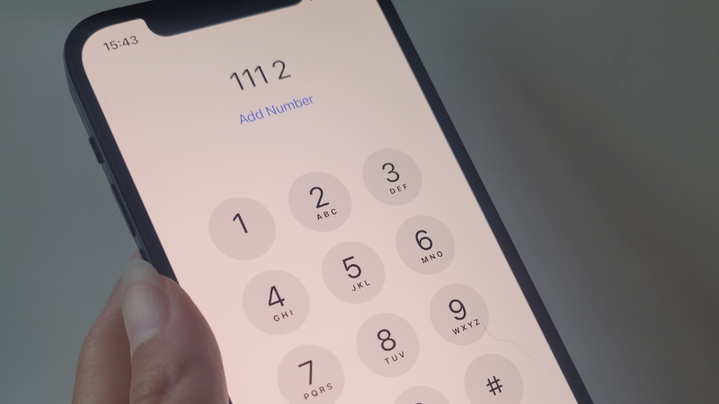 A person holding a smartphone with a dial pad displayed on the screen, showing the digits from 1 to 9, an asterisk, and hash symbol. The phone displays "Add Number" beneath a dialled number of '111 2' at the top, with the time 15:43.