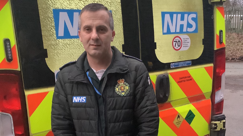 Dan - a man with short hair in a green uniform stood infront of an ambulance