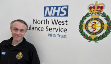 Person in a black North West Ambulance Service NHS Trust uniform smiling and standing in front of a wall with the NHS lozenge and North West Ambulance Service logo.