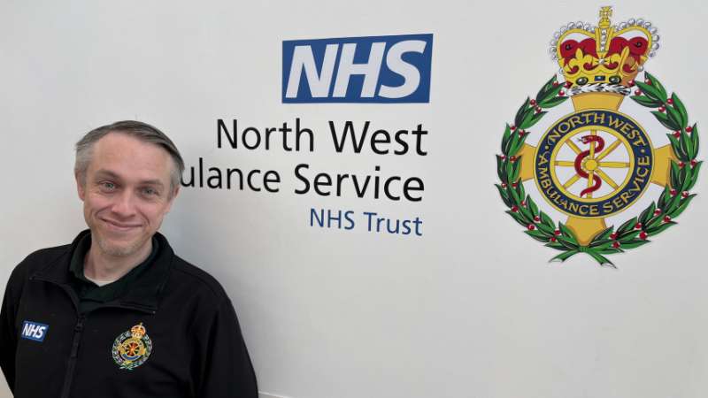 Person in a black North West Ambulance Service NHS Trust uniform smiling and standing in front of a wall with the NHS lozenge and North West Ambulance Service logo.