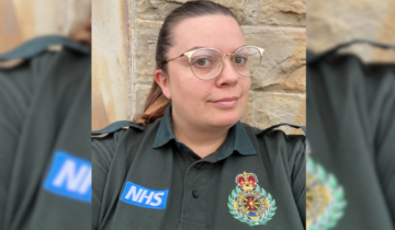 Laura in her green ambulance service uniform, featuring a badge with the service emblem and NHS logo, posing in front of a stone wall