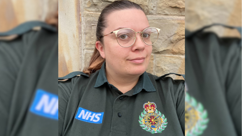 Laura in her green ambulance service uniform, featuring a badge with the service emblem and NHS logo, posing in front of a stone wall