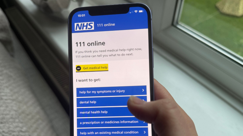 Hand holding a smartphone displaying the NHS 111 online help menu with options for medical assistance.