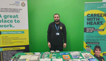 Jaffa in NWAS uniform standing at a NWAS careers fair stand with various banners and promotional materials.