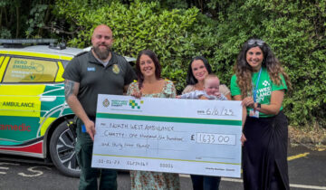 Four people and baby holding a large charity cheque. The group are standing in front of an emergency vehicle.