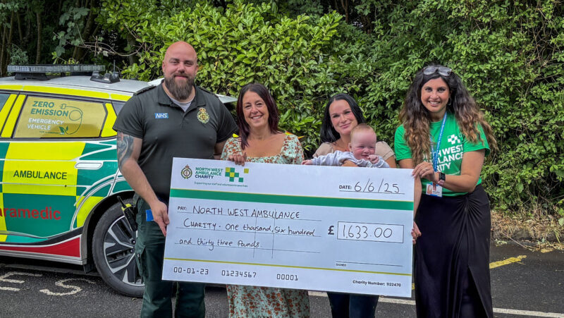 Four people and baby holding a large charity cheque. The group are standing in front of an emergency vehicle.