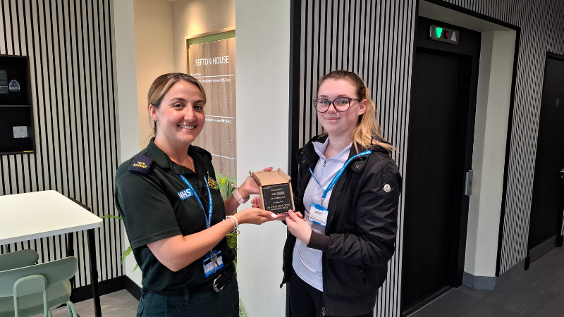 Two NHS 111 staff members holding a plaque between them, smiling at the camera.