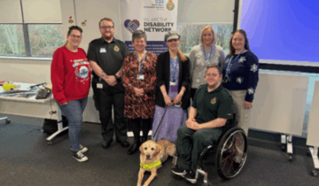 Seven members of our staff Disability Network, stood in front of a network promotional banner smiling to camera. Network Co-Chair Francesca is stood fourth-from-left with her guide dog.