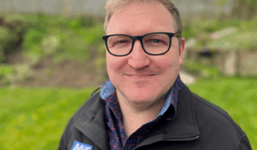 Mike, wearing an NHS jacket with an emblem, standing outdoors with greenery in the background.