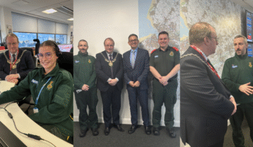 An office setting with several people interacting. One person is seated at a desk with computer screens displaying various data. Others, dressed in business attire and uniforms, are standing together. A large map and monitors showing information are visible in the background.