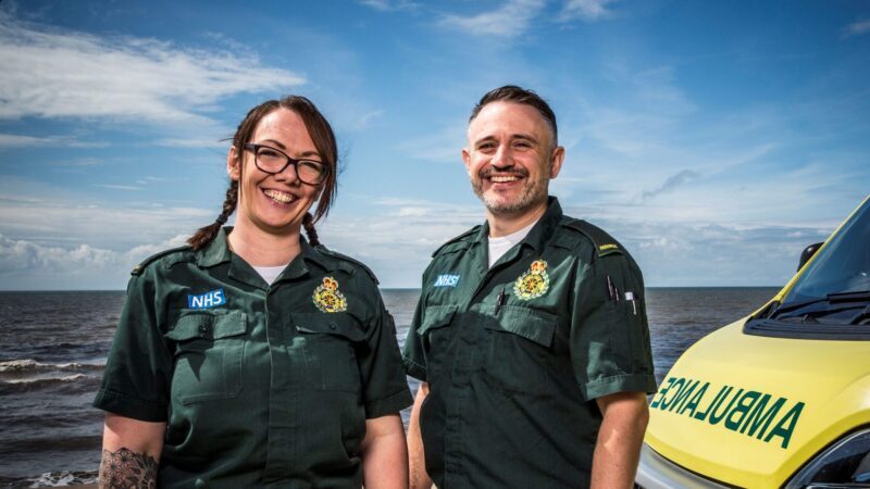 A man and a woman smiling. The right you see the bonnet of an ambulance