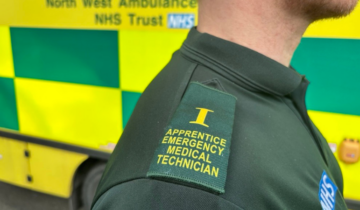 An individual wearing a green ambulance uniform with an epaulette reading "Apprentice Emergency Medical Technician" stands near an NHS ambulance.