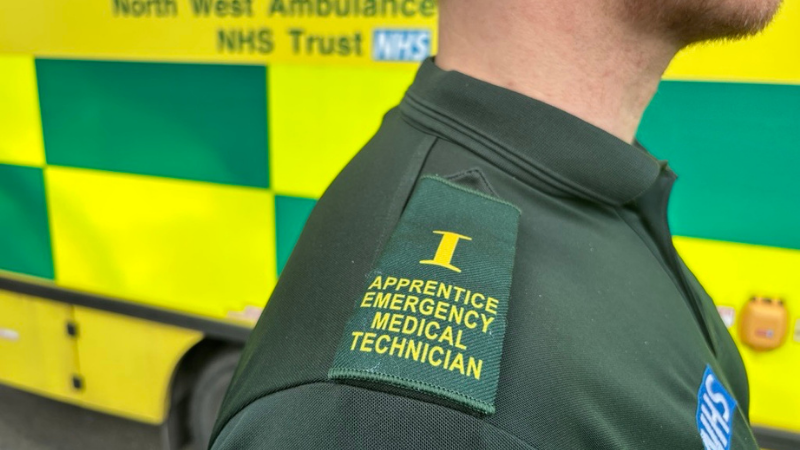 An individual wearing a green ambulance uniform with an epaulette reading "Apprentice Emergency Medical Technician" stands near an NHS ambulance.