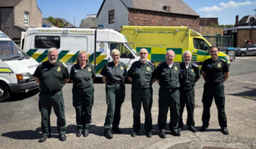 Seven people stood in front of a row of a mixture of modern and vintage ambulances