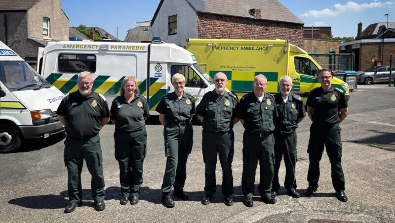 Seven people stood in front of a row of a mixture of modern and vintage ambulances