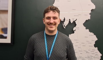 Rhys is standing in front of a white map mounted on a wall. He is smiling and wearing a jumper and a lanyard.