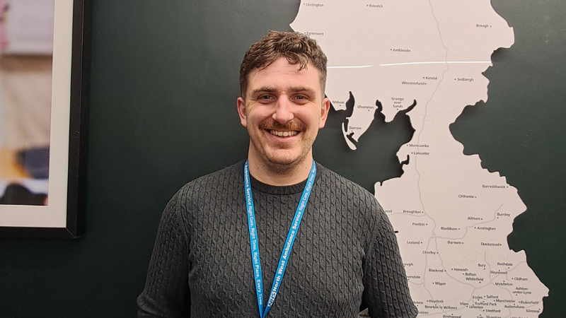 Rhys is standing in front of a white map mounted on a wall. He is smiling and wearing a jumper and a lanyard.