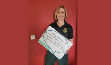 Maxine, in uniform holding a Freedom to Speak Up board that says, "I pledge to always be available to listen in a non-judgemental way and to support and encourage the person speaking up." She is stood in front of a red wall.