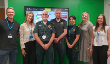seven people, some in uniform, stood together smiling, There is a green wall with a TV screen behind them.