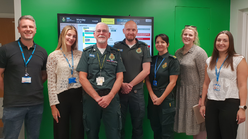 seven people, some in uniform, stood together smiling, There is a green wall with a TV screen behind them.