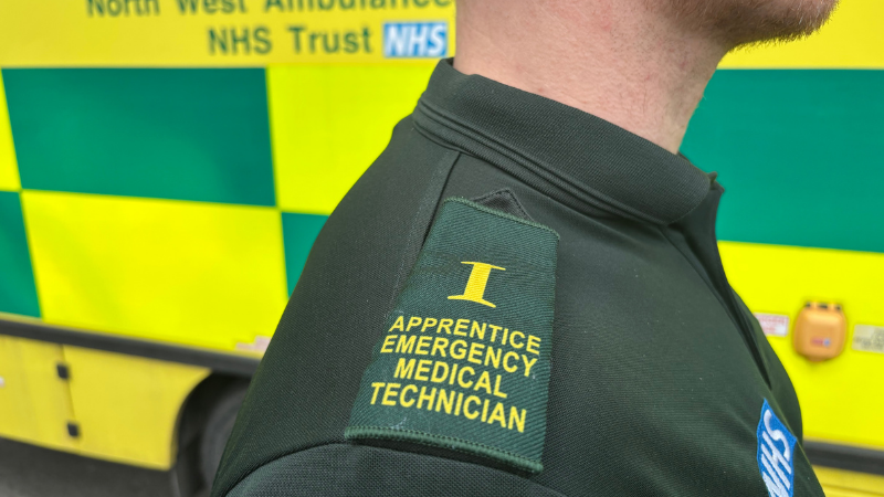 A person wearing a uniform with an epaulette reading "Apprentice Emergency Medical Technician" is standing next to a North West Ambulance NHS Trust vehicle.