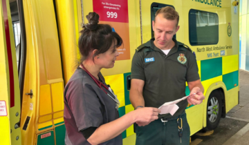 Merit doctor talking with a paramedic with an ambulance behind them. They are holding a piece of paper between them and engaged in discussion.