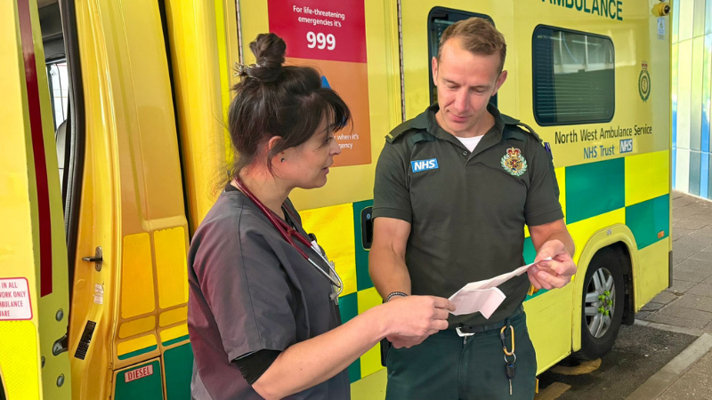 Merit doctor talking with a paramedic with an ambulance behind them. They are holding a piece of paper between them and engaged in discussion.
