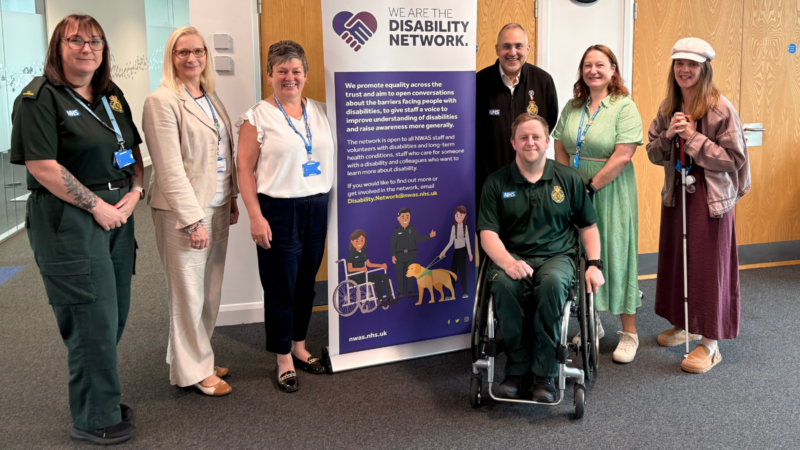 A group of seven people, some in uniform, others in office attire, posing in front or by the side of a disability network banner