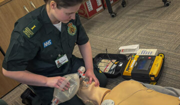 Woman wearing green ambulance uniform with apprentice medical technician written on her epaulette, is placing a resuscitation mask over a mannequin.
